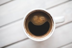 A bird's-eye-view of a cup of coffee with the foam in a form of a smiley face, in a white cup, on top of a white, wooden table.