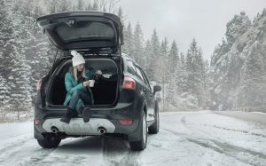 A woman with a white snow hat and green coat is sitting in her SUVs trunk with the door propped open and she is enjoying a hot drink on a snowy day.