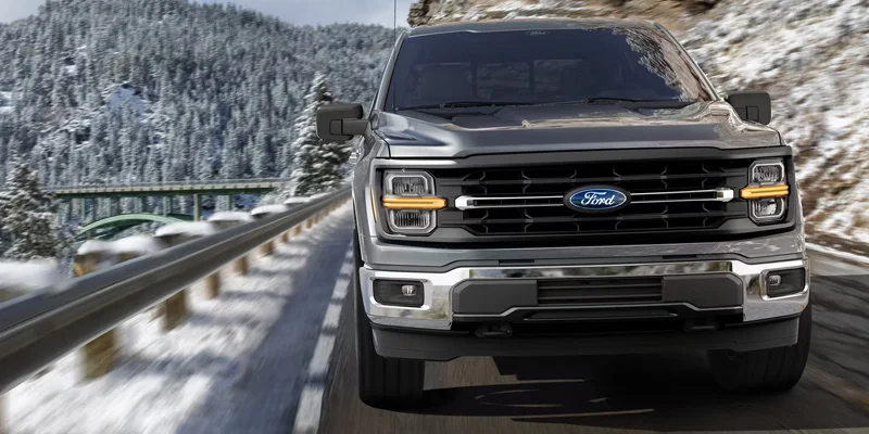 A front view of a Ford F-150 truck driving on a snowy road with a mountainous winter landscape in the background.